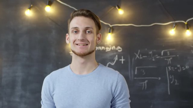 Handheld medium shot of young student or teacher standing at blackboard with formulas written on it, and looking at camera. Suitable for deepfake generation or face swap