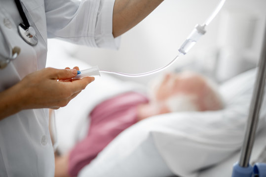 Medical Treatment. Close Up Of Female Arm Regulating IV Infusion By Using A Roller Clamp. Old Man Lying In Bed On Blurred Background