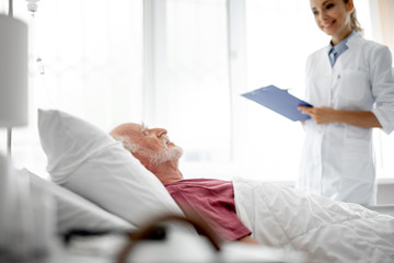 Obraz premium Side view portrait of old man resting in hospital room while young lady in white lab coat holding clipboard. Focus on bearded gentleman