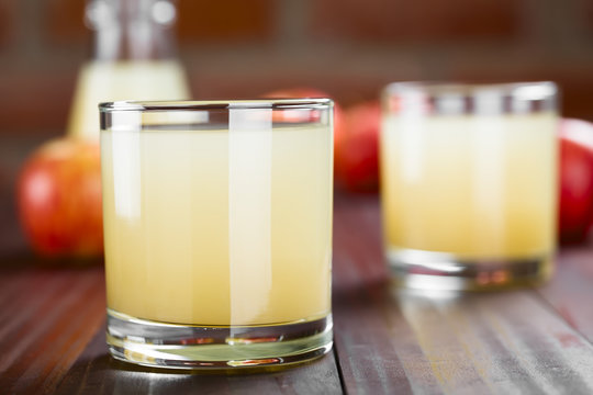 Refreshing apple juice in glasses with apples and bottle in the back, photographed on wood (Selective Focus, Focus on the front of the glass rim)