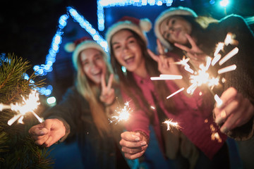 Selective focus of sparklers in hands of cheerful young smiling girls enjoying holiday time outdoors