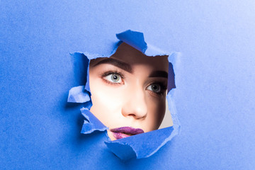 The face of a young beautiful girl with a bright make-up and puffy purple lips peers into a hole in...