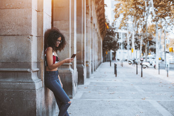 Young curly hipster girl in glasses is leaning against a stone pillar outdoors, smiling and typing message for her friend, with a copy space zone on the right for your logo or a text advert message