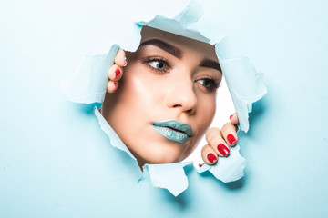 Young woman with beautiful bright eyes with brown shadows, lipstick of the blue color and eyebrows looks into the hole of the colored paper.
