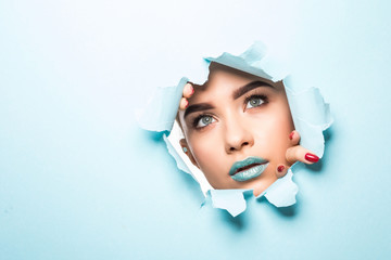 Face of a young beautiful girl with a bright make-up and puffy blue lips peers into a hole in blue paper.