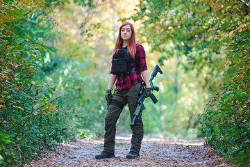 Woman with a rifle on the road in forest/Pretty woman with a gun in her hands and in a black vest stands in the middle of the road in the forest and looks into the camera