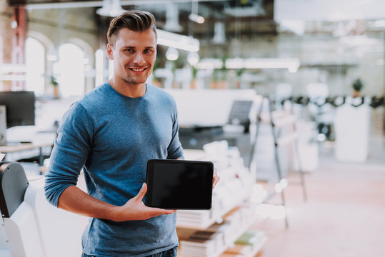 Waist up portrait of smiling guy standing in modern office and holding tablet in hands. Copy space in right side