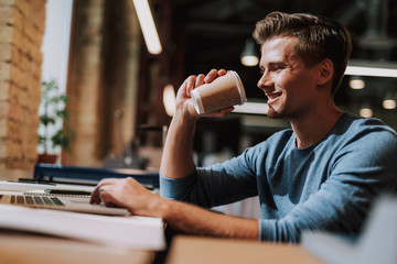 Waist up portrait of smiling man sitting in coworking space and working with laptop while holding...