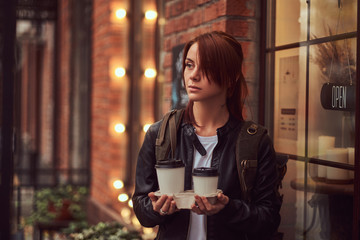 A charming girl wearing a leather jacket with a rucksack holding cup with takeaway coffee outside near the cafe.