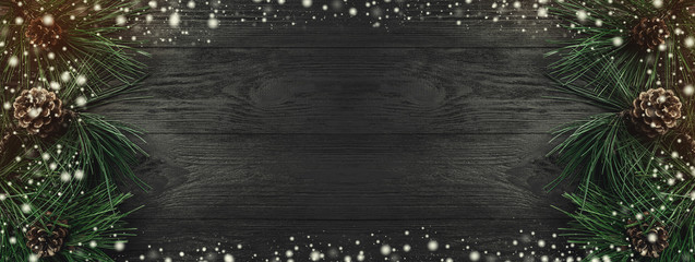 Christmas card. Black wood background, with pine branches and pine cones from one side, top view. Xmas rectangle congratulation. Effect of light and snow.