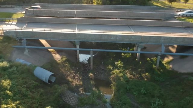 Aerial crane down shot of light traffic driving over a man made drainage canal or ravine crossover bridge on a sunny afternoon.