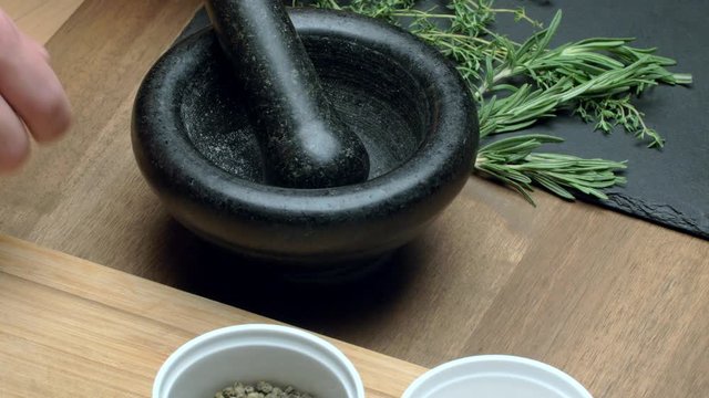 Person's hands putting black peppercorns into a dark stone mortar and pestle for crushing, with herbs and spices laid out on a wooden table