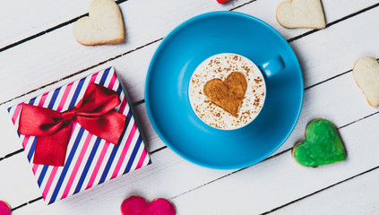 Cup of Cappuccino with heart shape symbol and cookies with gift box on white wooden background