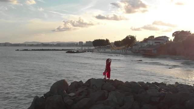 Aerial footage of the Young beautiful woman in long red dress on a mountain cliff watching at sea, young girl in red dress with hair waving in the wind at the sea on the rock