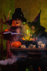 Image of laughing two witches in black hats reading book of incantations on cauldron with steam