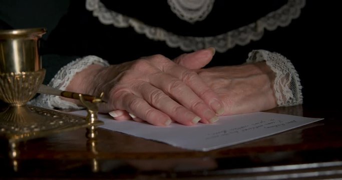 An ornate inkwell with a holder for its fancy dip pen and the folded hands of an old Victorian era woman with an unfinished letter who then exits the scene.
