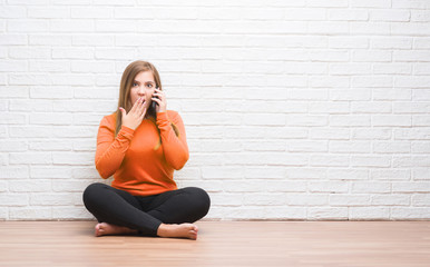 Young adult woman sitting on the floor over white brick wall calling using smartphone cover mouth with hand shocked with shame for mistake, expression of fear, scared in silence, secret concept