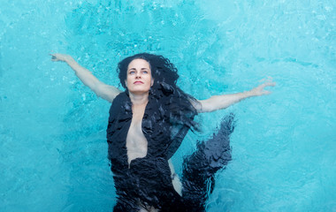 schöne erwachsene Frau mit dunkel braunen lockigen Haaren in schwarzem Tuch, die Arme seitwärts ausgestreckt, glücklich schwimmend im Wasser im  Pool, türkisblau schimmert das kristallklare Wa