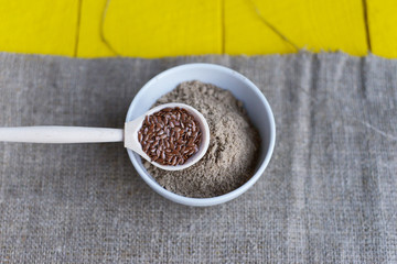 bowl of flax flour, a spoon with flax seeds on a yellow wooden background and linen canvas