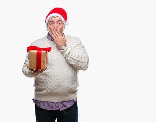 Handsome senior man wearing christmas hat and holding gift over isolated background cover mouth with hand shocked with shame for mistake, expression of fear, scared in silence, secret concept