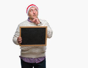Handsome senior man wearing christmas hat and holding blackboard over isolated background serious...