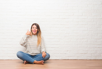 Fototapeta na wymiar Young adult woman sitting on the floor over white brick wall at home smiling doing phone gesture with hand and fingers like talking on the telephone. Communicating concepts.