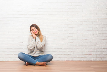 Young adult woman sitting on the floor over white brick wall at home sleeping tired dreaming and posing with hands together while smiling with closed eyes.