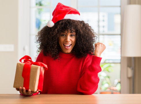 African american woman holding present wearing christmas red hat screaming proud and celebrating victory and success very excited, cheering emotion