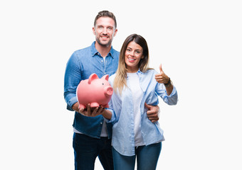 Young couple in love holding piggy bank over isolated background happy with big smile doing ok sign, thumb up with fingers, excellent sign