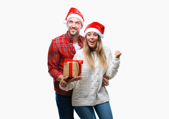 Young couple in love wearing christmas hat and holding present over isolated background screaming proud and celebrating victory and success very excited, cheering emotion