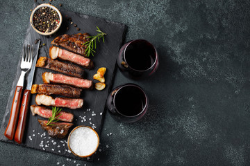 Obraz na płótnie Canvas Sliced steak Striploin, grilled with pepper, garlic, salt and thyme served on a slate chopping Board with a glass of red wine on a dark stone background. Top view with copy space