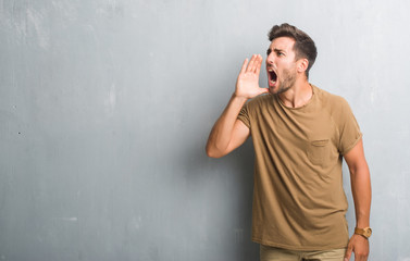 Handsome young man over grey grunge wall shouting and screaming loud to side with hand on mouth. Communication concept.