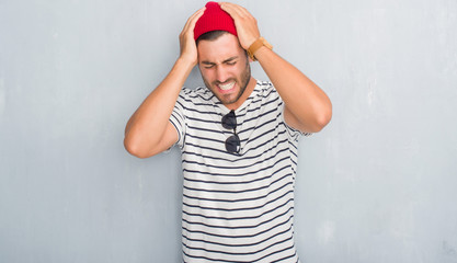Naklejka premium Handsome young hipster man over grey grunge wall wearing navy t-shirt and wool cap suffering from headache desperate and stressed because pain and migraine. Hands on head.
