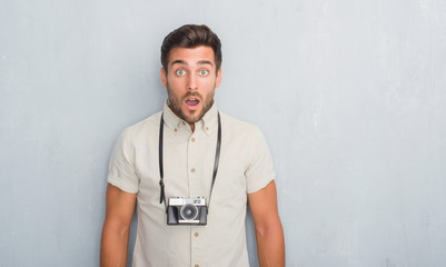 Handsome young man over grey grunge wall holding vintage photo camera scared in shock with a...