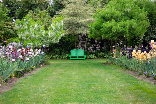 Elevated level perspective of English style garden path of mowed lawn, mixed flower beds, trees, and green adirondack garden bench, daytime, no sky
