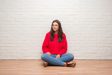 Young brunette woman sitting on the floor over white brick wall with a confident expression on smart face thinking serious