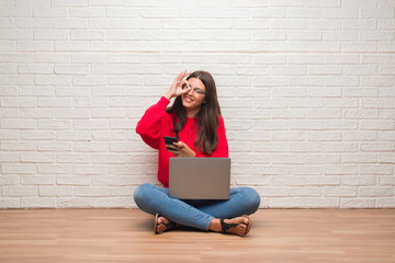 Young brunette woman sitting on the floor using laptop and smartphone with happy face smiling doing ok sign with hand on eye looking through fingers