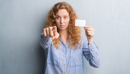 Young redhead woman over grey grunge wall holding blank visit card pointing with finger to the...