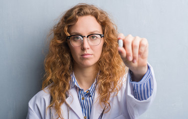 Young redhead doctor woman over grey grunge wall holding a pill with a confident expression on smart face thinking serious