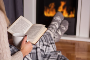 Girl in front of the fireplace reading book and warming feet on fire and legs are covered with blanket, in hand holds cup of hot tea. Winter and cold weather concept at home. Close up, selective focus