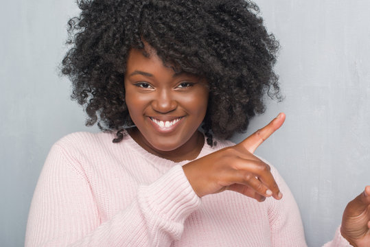 Young african american plus size woman over grey grunge wall wearing winter sweater smiling and looking at the camera pointing with two hands and fingers to the side. - Powered by Adobe