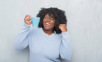 Young african american woman over grey grunge wall drinking a cup of coffee screaming proud and celebrating victory and success very excited, cheering emotion