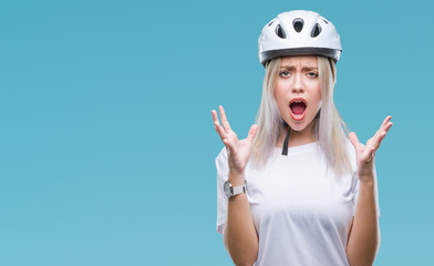 Young blonde woman wearing cyclist security helmet over isolated background crazy and mad shouting and yelling with aggressive expression and arms raised. Frustration concept.