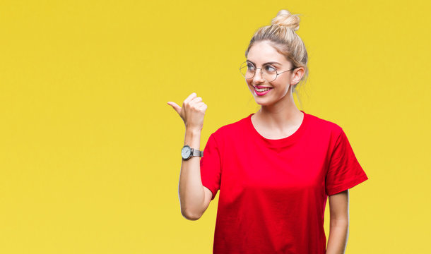 Young beautiful blonde woman wearing red t-shirt and glasses over isolated background smiling with happy face looking and pointing to the side with thumb up.