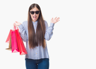 Young Chinese woman over isolated background holding shopping bags on sales very happy and excited, winner expression celebrating victory screaming with big smile and raised hands