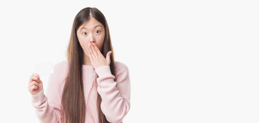 Young Chinese woman over isolated background holding visit card cover mouth with hand shocked with shame for mistake, expression of fear, scared in silence, secret concept