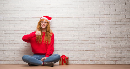 Young redhead woman sitting over brick wall wearing christmas hat doing happy thumbs up gesture with hand. Approving expression looking at the camera with showing success.