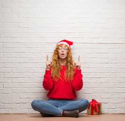 Young redhead woman sitting over brick wall wearing christmas hat amazed and surprised looking up and pointing with fingers and raised arms.