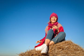 Naklejka na ściany i meble Portrait of a beautiful young model in knitted hat and warm clothes enjoy day, on background haystacks in sunny autumn day . Autumn warm photo. Woman smiling and look away, joyful cheerful mood.