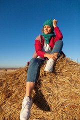 Naklejka na ściany i meble The concept of livestyle outdoor in autumn. Close up of a young woman student in a warm autumn clothes looking funny, smilling, posing on the haystack